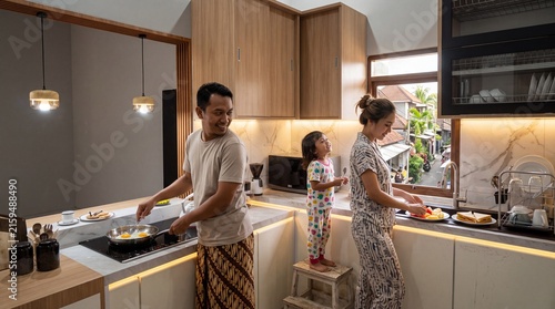 Happy young family with a small child preparing a healthy breakfast in a bright modern kitchen with wooden cabinets and warm morning light