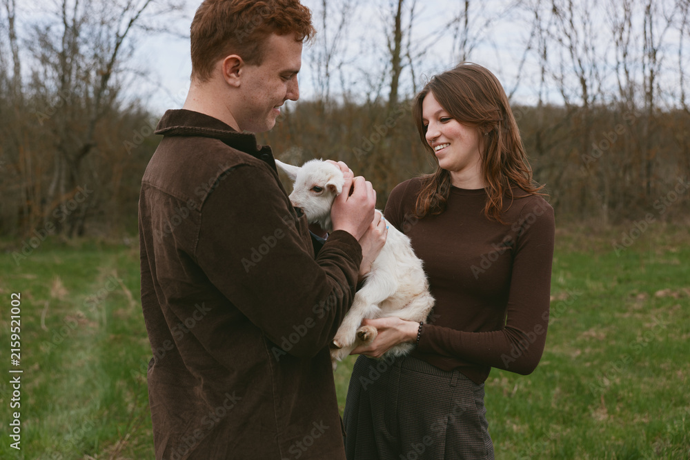 Naklejka premium Casual couple outdoors gently care for a white goat kid, sharing a tender moment in a rustic field, soft natural light, warm sweaters, calm smile, and peaceful, affectionate mood