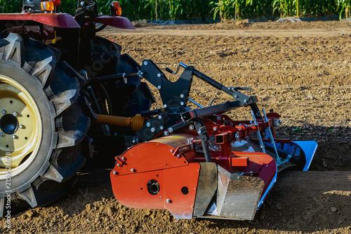 Принти на полотні Agricultural tractor standing with front blade and rear rotary tiller on tilled agricultural field during land preparation for farming with trees and cornfield in background