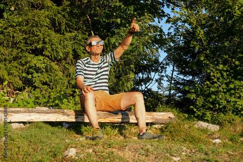 A man sitting on a wooden bench looks directly up at the sky through special solar eclipse glasses. He points with his index finger at the astronomical event of the solar eclipse