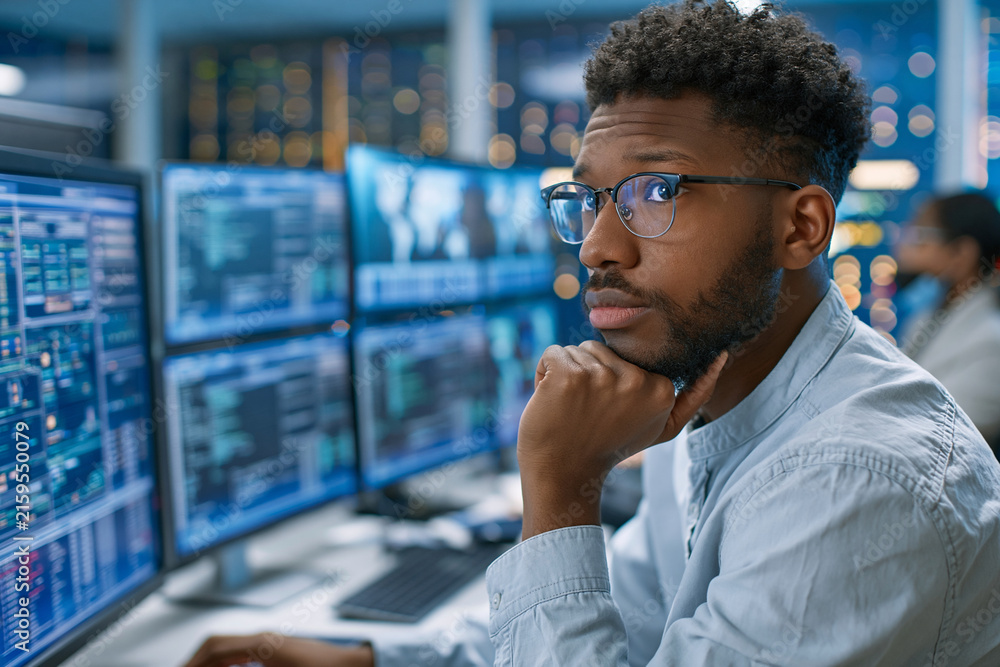 Fototapeta premium Focused analyst studies complex data on multiple screens. Dark glasses, thoughtful gaze, hand resting near chin. Blue-lit workspace with glowing monitors displaying charts