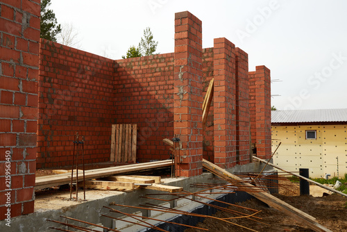 Brick house under construction with load-bearing walls, reinforced concrete foundation, and unfinished structural columns.