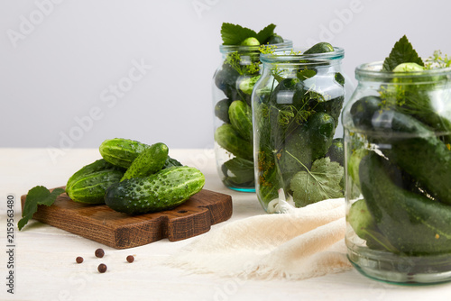  Cucumbers in glass jars and on a cutting board, dill, garlic, and spices on a white wooden background.