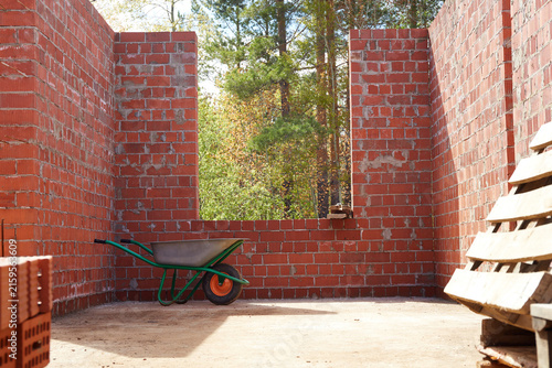 Interior of an unfinished brick house with a wheelbarrow standing near a large window opening