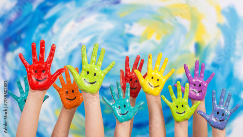 Childrens hands painted in bright multicolored poster paints with smiling face details are held up together in front of a blue and yellow painted artistic background.