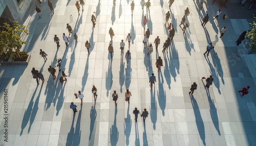 A crowd of people walk across a tiled plaza casting long shadows in the sunlight.