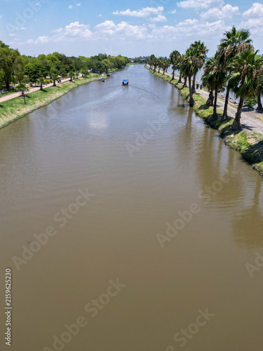 An aerial perspective captures the tranquil canal in Jamay, Jalisco. A small boat glides along the waterway, flanked by lush green trees and stately palm trees under a bright blue sky. Idyllic landsca