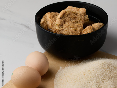 Raw soy cutlets soaking in aromatic vegetable broth, placed in a black bowl and ready for breading. Fresh chicken eggs and fine breadcrumbs on parchment paper are visible nearby. Ingredients for a veg