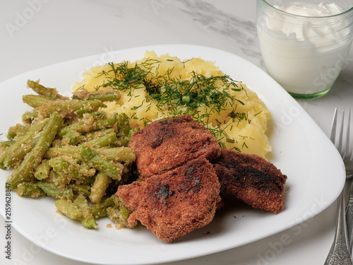 Homemade vegetarian dinner with fried soy cutlets, mashed potatoes sprinkled with fresh dill, and green beans with breadcrumbs, served on a white plate next to a glass of sour cream and a fork.