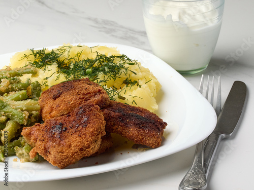 Homemade vegetarian dinner with fried soy cutlets, mashed potatoes sprinkled with fresh dill, and green beans with breadcrumbs, served on a white plate next to a glass of sour cream and a fork.
