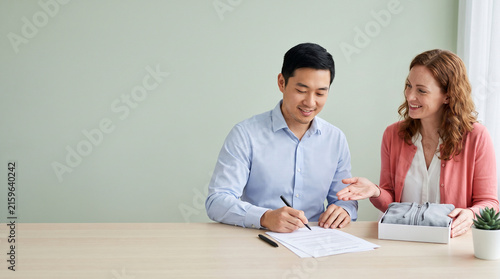 Asian man signing contract document with female client or consultant, business entrepreneur sealing deal or franchise agreement at office desk with clothing product sample banner
