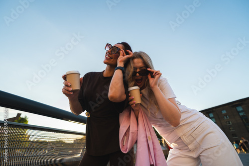 Photography Two cheerful female friends holding coffee cups and smiling on an urban bridge