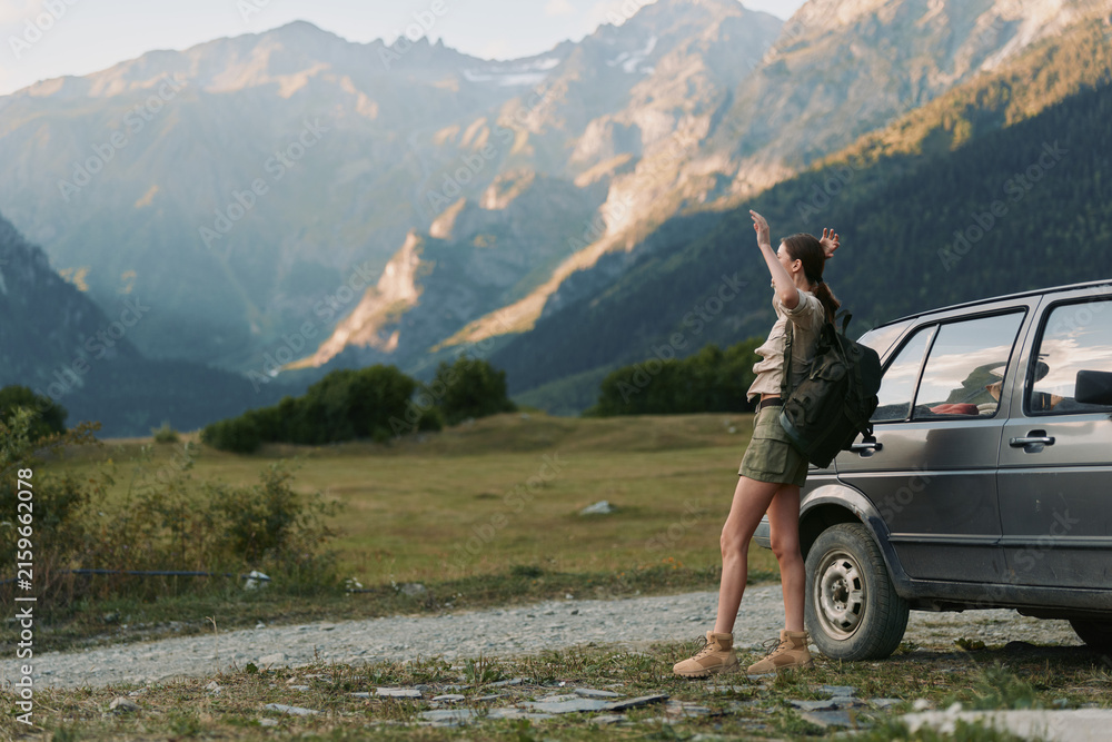Naklejka premium Woman with backpack standing near car on mountain road pointing at distant peaks. Outdoor lifestyle, adventure, travel, and nature exploration concept in warm daylight.