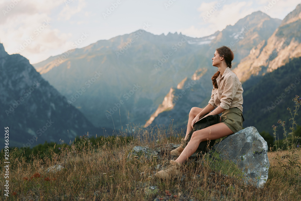 Fototapeta premium A woman in casual clothes sits on a rock in a mountain meadow, looking at the distant peaks during a calm sunset. Concept for lifestyle, nature, and relaxation.