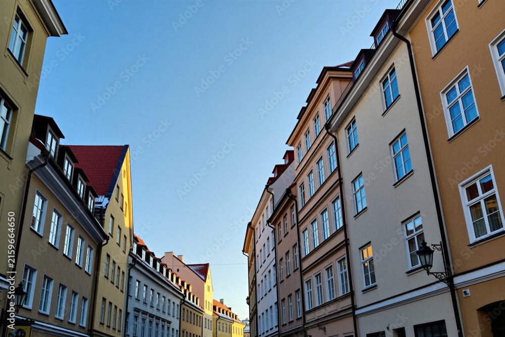 Obraz premium Low angle view of narrow city street with tall historical buildings on clear day. Sunlight illuminates colorful house facades under blue sky. Old town architecture in Europe stands along alleyway.