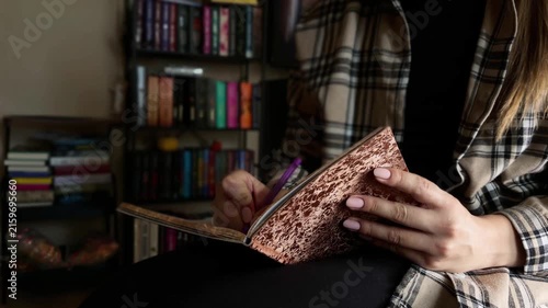 Woman's hands writing notes in a diary journal with a pen. Unrecognizable female taking notes in a notebook while sitting in a library next to a bookshelf at home