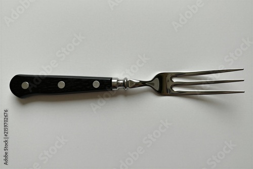 Metal carving fork with black wooden handle rests on plain white surface. Stainless steel utensil features three sharp tines for piercing large food cuts. Classic culinary tool design offers pro grip