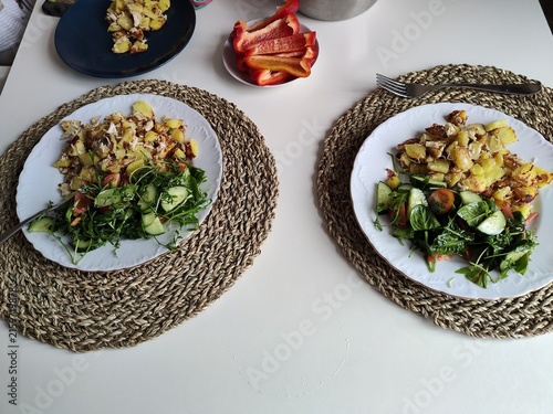 Obraz na plátně Two plates of fresh salad with leafy greens, cucumber, tomatoes, and sliced vegetables served on woven placemats, healthy homemade meal, and food photography
