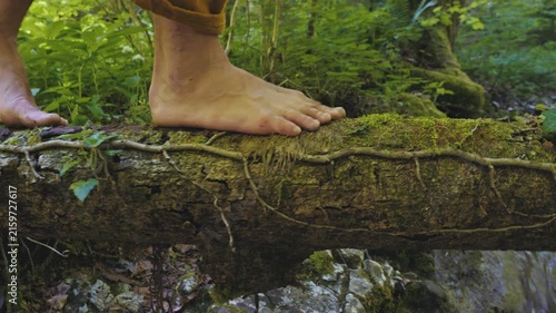 A person practices a zen-like balance exercise on a moss-covered log in a serene forest. Bare feet focus on maintaining stability, surrounded by lush greenery and a flowing stream.