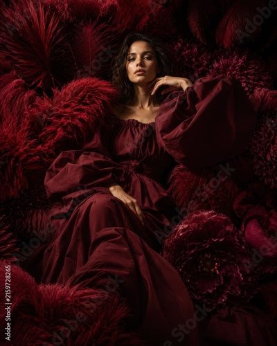 Surreal fine art portrait of an alluring woman in a burgundy puff sleeve gown surrounded by deep red exotic feathers and giant velvet flowers in moody dramatic chiaroscuro lighting