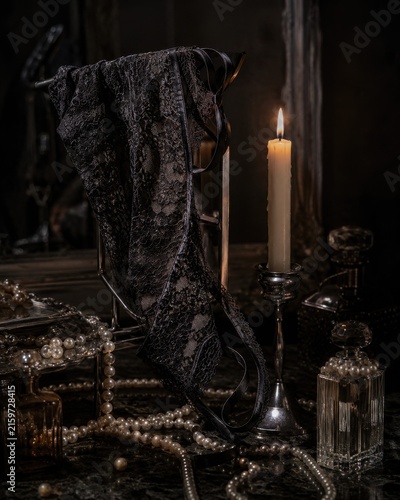 Dark moody still life with black lace lingerie draped over a metal stand beside a burning candle, shimmering pearls, and vintage perfume bottles on a dark table
