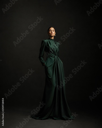 Regal full length portrait of an aristocratic brunette woman in an emerald green silk gown with structured shoulders standing in low key theatrical lighting against dark background