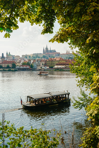 Canvas Print Barco turístico navegando por el río Moldava con vistas al castillo de Praga y el centro histórico rodeado de vegetación verde