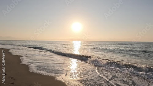 Slow motion video walking along the brown sand of the beach shore watching the sun at sunrise or sunset over the horizon of the calm Mediterranean Sea, with a clear sky
