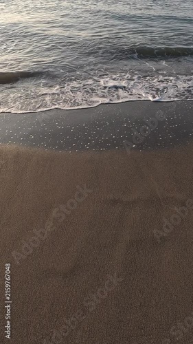 Vertical video close-up view of ocean waves gently rolling onto a sandy beach. The foamy white water contrasts with the brown wet sand as the tide ebbs and flows along the shoreline
