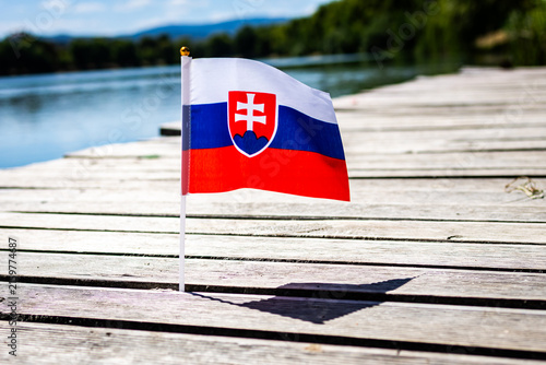Photography Slovak flag with coat of arms and red white blue bands on a slim pole beside a lake