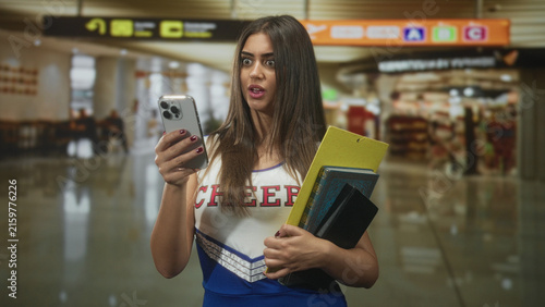 Quadro su tela Woman cheerleader in blue uniform holds phone and notebooks in airport terminal, reading a message with a surprised, puzzled expression; surprise uncertainty