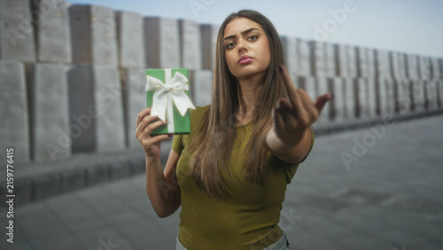 Fotografie Young woman holds green gift and shows middle finger on street with a stern expression; defiance rejection
