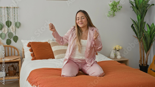 Konstfotografi Woman kneeling on bed in pink pyjamas stretches arms and smiles in a bedroom building; morning joy relaxation