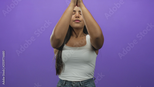 Bild auf Leinwand Young woman pressing hands together in prayer at chest, eyes closed, wearing white tanktop and jeans in a purple studio; calm reflection