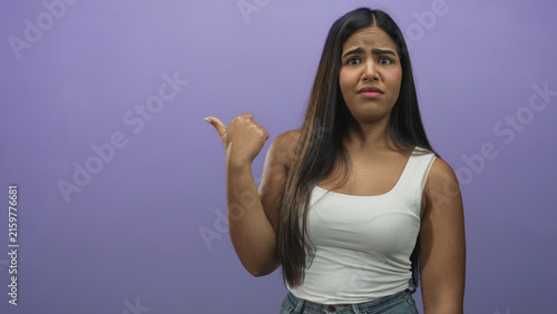 Obraz na plátně Young hispanic woman pointing thumb over shoulder in purple studio wearing white tank top; skepticism hesitation