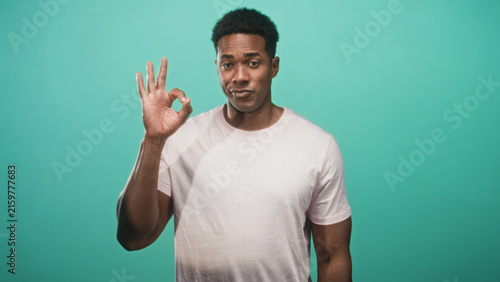 Fotografie Man in pale pink t shirt making ok sign with right hand while facing camera against teal backdrop in studio; playful approval
