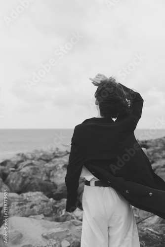 Принти на полотні Wind in coat over rocky coast, silhouette facing sea, black and white scene capt