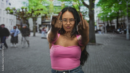 Canvastavla Woman young hispanic brunette shows middle fingers to camera on busy city street wearing pink top and glasses; defiance