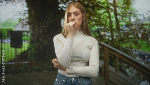 Фотография Woman with hand to mouth and crossed arm by wooden railing on park walkway wearing cream sweater and jeans; thoughtful solitude