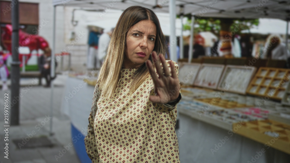Fototapeta premium Woman with palm raised in a stop gesture by a street market stall, patterned blouse and rings visible, direct serious gaze; defiance.