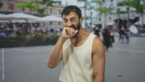 Obraz na plátne Man biting his fist, hand to mouth gesture in a busy street cafe wearing a sleeveless tanktop, bearded middleaged hispanic; stifled amusement
