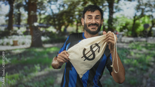 Принти на полотні Man in soccer jersey holding a money bag with dollar sign, smiling and inspecting its drawstring while standing in forest; satisfaction