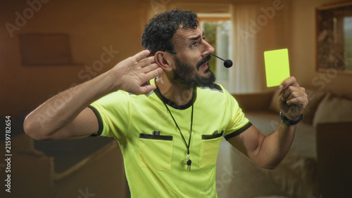 Obraz na plátne Man referee holding a yellow card and cupping his ear inside a building, wearing a whistle and headset; official authority alert