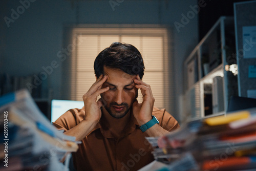 Stressed young Latin businessman having severe headache and migraine at desk in night office. Debt crisis, corporate burnout, late overtime workload, mental health anxiety, business failure concept.