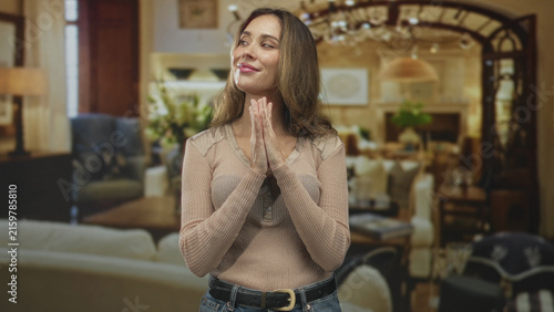 Fotografie Woman clasping hands near chest smiling in a knit sweater and jeans, posed in a warm building interior with sofa and chandelier; hopeful calm