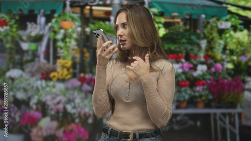 фотографія Woman speaking into smartphone, holding phone close to mouth with hand raised in a questioning gesture at a street flower market; concern communication request