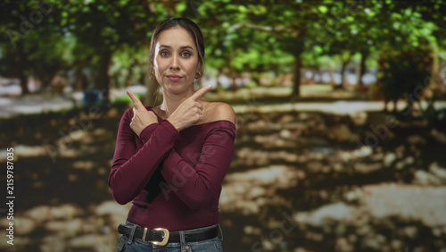 Fotografía Woman points index fingers to opposite sides with crossed arms, wearing burgundy sweater and jeans, standing before trees in forest; indecision choice