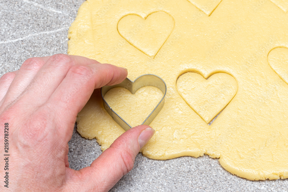 Obraz premium Pastry chef's hand cuts heart-shaped cookies from raw yellow dough on a gray table background, close-up.