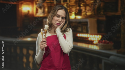 Foto Woman in red apron holds makeup brushes and presses cheek with hand, grimacing in an ornate building with candlelit altar; toothache pain
