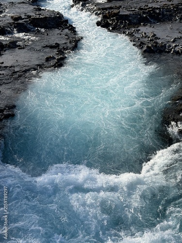 Blaskogabygg, Iceland - May 08, 2026: Turquoise water flows through rocky terrain, creating a natural stream with foamy waves and textured black stones along the banks